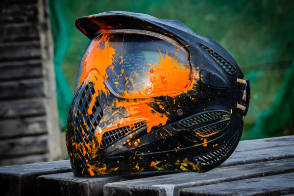 A Paintball Mask at the local Playing Field Sitting On a Table Splattered With Orange Paint after Being Used in a Large Game and clearly been hit several times by multiple headshots